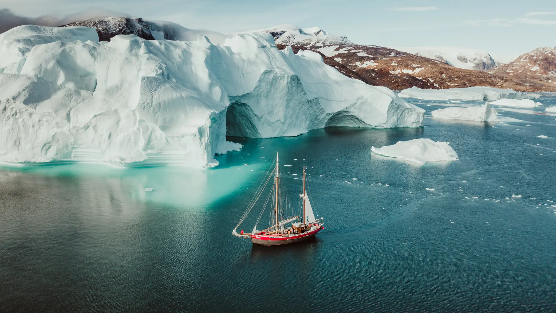 A red two-masted sailing vessel crossing dark water beside a monumental Greenland iceberg with a glowing turquoise base
