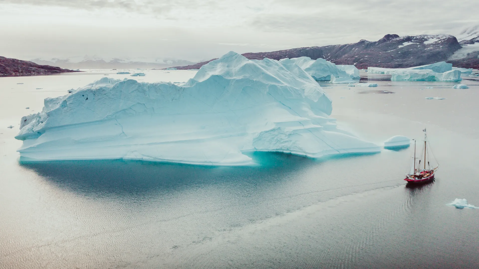 A small red sailing vessel navigating calm silver water past an immense pale-blue iceberg in East Greenland